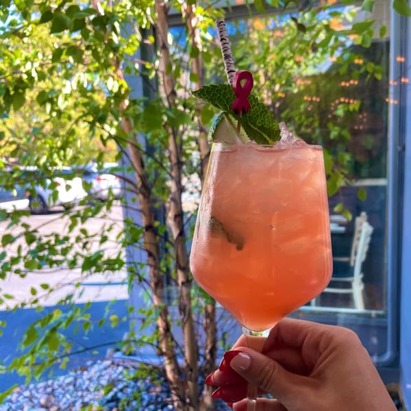 A hand holds a pink cocktail with mint and a red ribbon garnish, outdoors at E+O Kitchen.