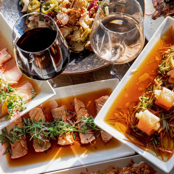 An overhead view of various Asian-fusion dishes and wine glasses on a wooden table.