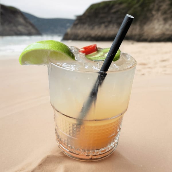 A refreshing cocktail with lime, chili, and a black straw on a sandy beach with ocean and cliffs.