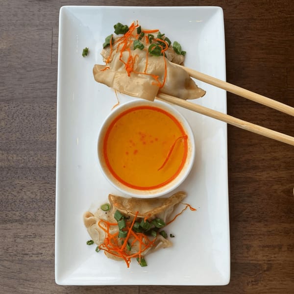 Chopsticks holding a dumpling over a bowl of orange sauce, with more dumplings on a white plate.