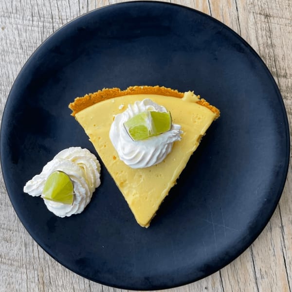 A slice of key lime pie with whipped cream and lime wedges on a dark plate.