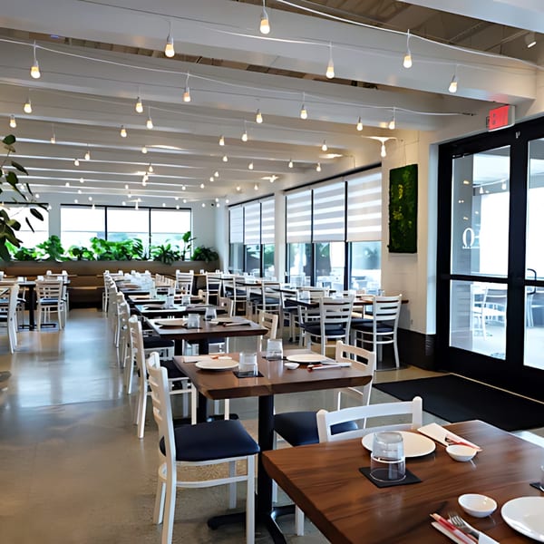 E+O Kitchen's spacious dining room with many tables set, large windows, and hanging lights.
